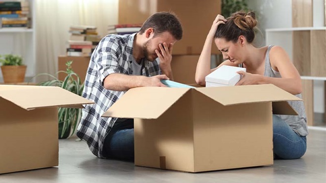 Casal com ar deprimido a arrumar objetos em caixas de cartão num ambiente doméstico rodeado de outras caixas abertas.