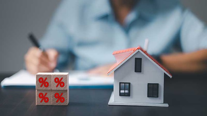 Casa e cubos de madeira com o símbolo de percentagem e um homem na retaguarda a escrever enquanto pondera a troca de casa