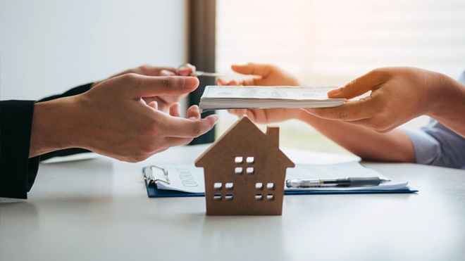Grande plano de umas mãos com documentos a passar a outra pessoa; maquete de pequena casa à frente, sobre uma mesa 