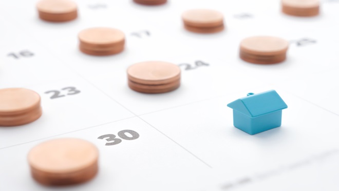 Imagem de página de calendário (mês) com moedas em cada dia e no último dia uma casa azul pequena - alusão ao pagamento de prestação de crédito à habitação