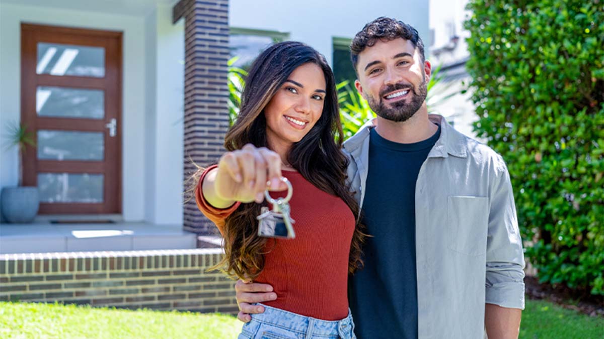 Duas pessoas em frente a uma casa moderna; uma está a apontar para a câmara e segura um molho de chaves, com a entrada e jardim ao fundo.