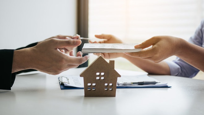 Grande plano de mãos a transferir documentos formais de uma para a outra, com uma casa em primeiro plano - alusão a transferência de crédito habitação