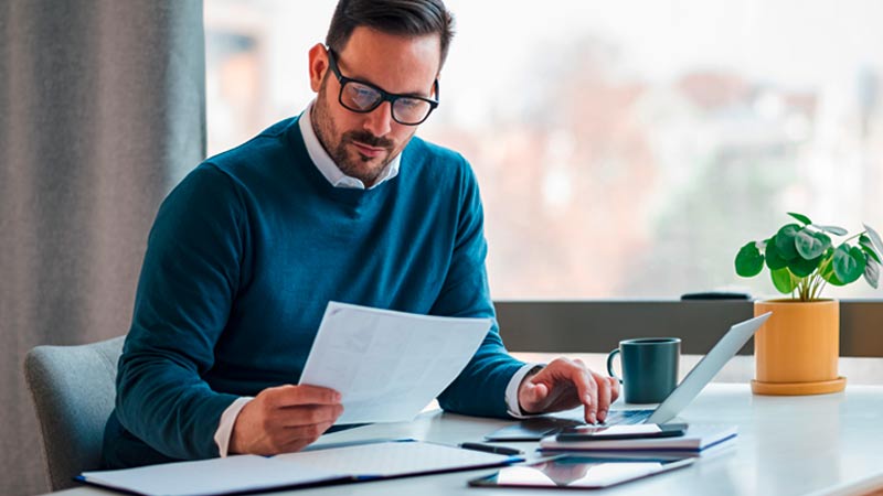Homem adulto sentado a uma secretária com um portátil, analisa documentos 