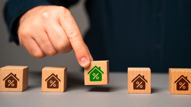 Grande plano de uma mesa com 5 cubos de madeira com símbolos de casa e percentagem; uma mão levanta o cubo central que tem o ícone a verde