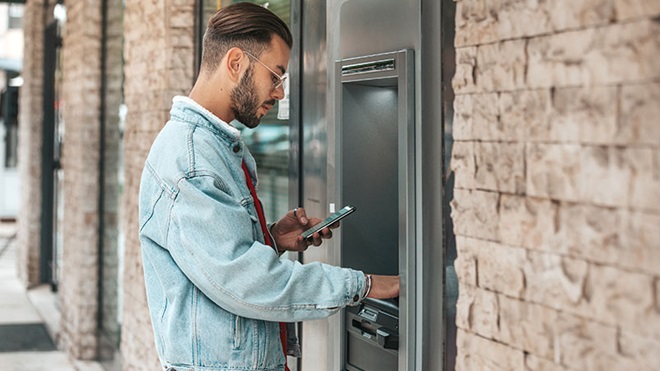 Homem utiliza caixa automática com a mão direita enquanto olha para telemóvel que segura com a mão esquerda