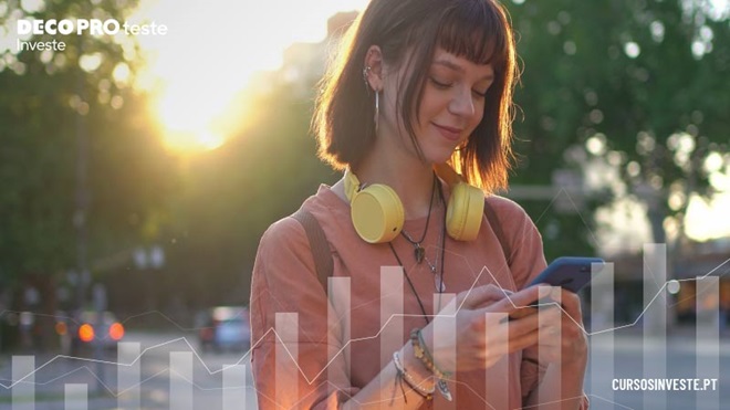 Mulher jovem na rua mexe no telemóvel, gráficos de barras com transparências à frente - alusão a cursos de literacia financeira da Proteste Investe
