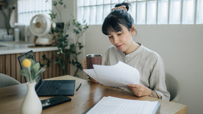 Mulher em casa a analisar documentos com um computador portátil à frente, sentada numa mesa