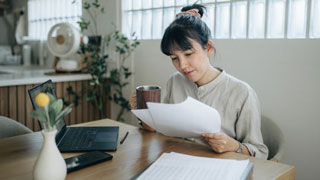 Mulher em casa a analisar documentos com um computador portátil à frente, sentada numa mesa