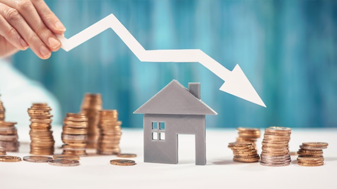 Grande plano de mão a segurar uma seta descendente por cima de uma casa: de um lado uma pilha de moedas grande; do outro (final da seta), uma pilha mais pequena. Alusão a decréscimo de pagamanento da prestação dacasa