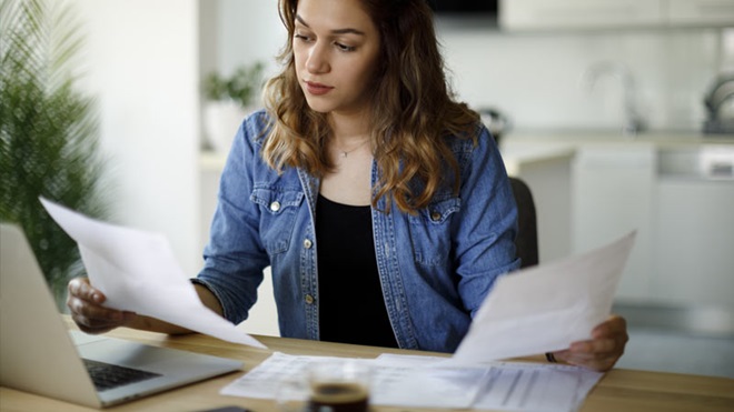 Mulher com ar confuso e dois documentos na mão, sentada em frente ao computador