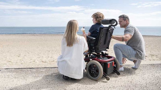 pai e mãe ao lado de criança em cadeira de rodas, de costas, a olhar para o mar