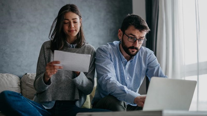 Pilha de moedas crescente numa mesa, ao lado de uma casa de madeira; uma mão vinda de cima coloca uma moeda na casa