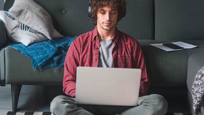 Jovem a preencher o IRS, sentado no chão da sala com um portátil e phones na cabeça, em frente a um sofá cinzento