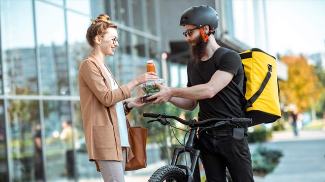 Estafeta de bicicleta e mochila a entregar comida a uma mulher à frente de uma empresa