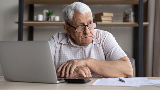 Senhor idoso, de óculos e cabelos brancos, sentado numa mesa com o portátil e documentos à frente, de ar pensativo