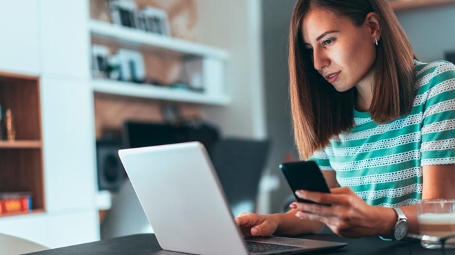 Jovem empresária sentada à secretária em frente de um portátil, com um telemóvel na mão, olha atentamente para os ecrãs