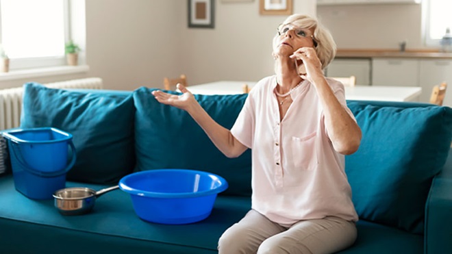 Mulher sentada num sofá azul ao telefone para acionar o seguro multirriscos-habitação, com balde e alguidar a apanhar a água que cai do teto