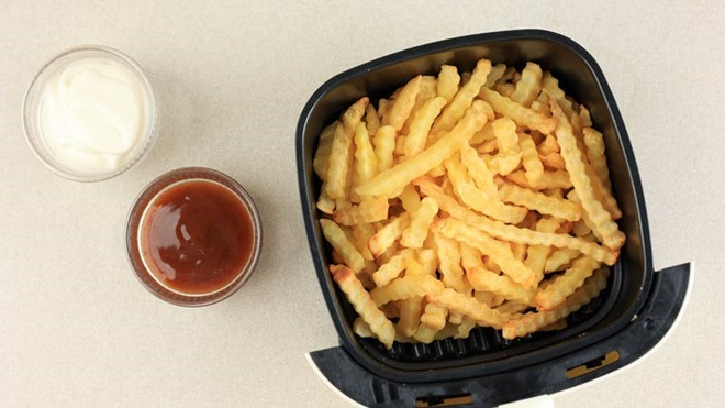 Grande plano visto de cima do cesto de uma airfryer cheio de batatas fritas douradas; duas taças com molhos ao lado