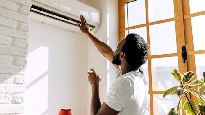 Homem segura um comando e aponta para ar condiconado, numa sala