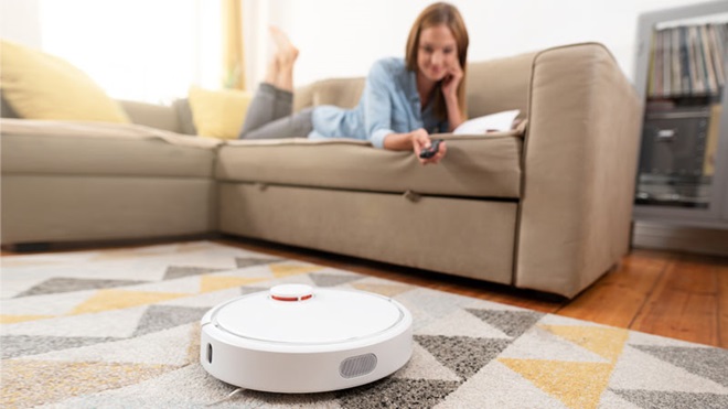 Mulher deitada no sofá com ar descontraído enquanto o arpirador robô aspira o tapete da sala
