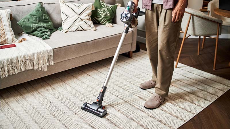 Pessoa a aspirar um tapete numa sala de estar, usando um aspirador vertical sem fios; sofá com almofadas