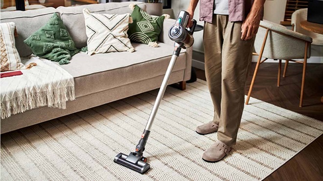Pessoa a aspirar um tapete numa sala de estar, usando um aspirador vertical sem fios; sofá com almofadas