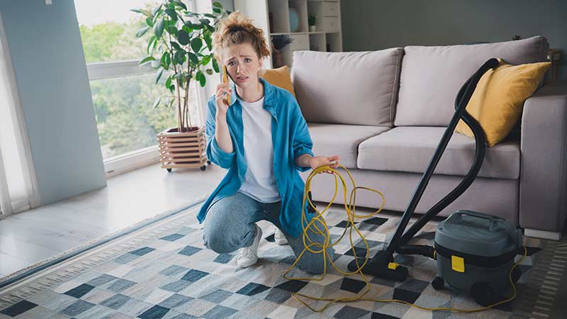 Mulher jovem numa sala de estar fala ao telemóvel com ar desesperado ao pé de aspirador vertical avariado