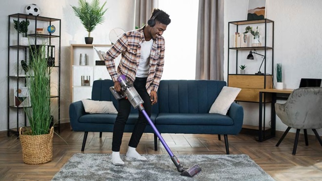 Um homem com auscultadores nos ouvidos aspira um tapete numa sala de estar com um aspirador vertical sem fio