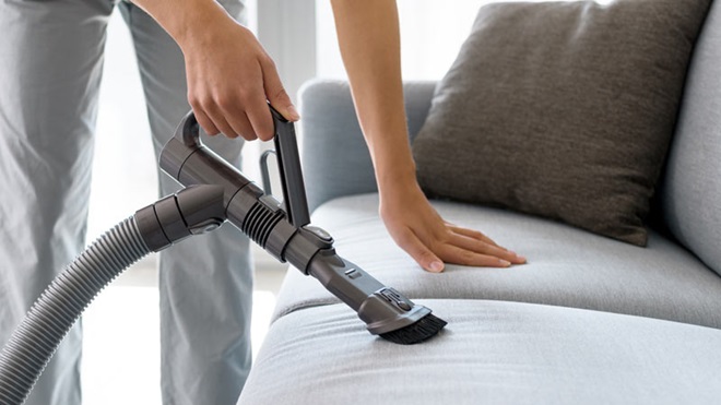 Uma pessoa tem a mão esquerda pousada num sofá cinzento enquanto com a mão direita aspira esse sofá