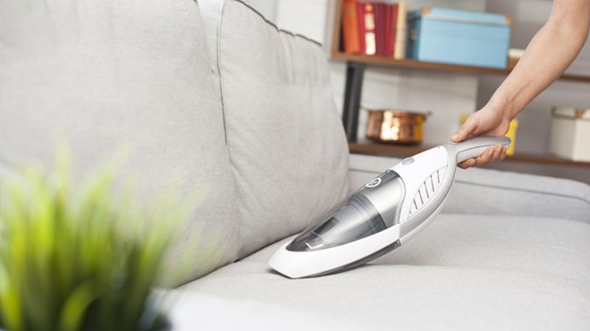 Numa sala de estar, uma mão segura num mini-aspirador para aspirar um sofá cinzento