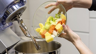 Plano de pormenor da parte superior de um robô de cozinha, com a tampa aberta, levantada. Do lado direito da imagem, um par de mãos agarra uma taça transparente contendo alguma fruta cortada, que se prepara para despejar para dentro do copo do robô.