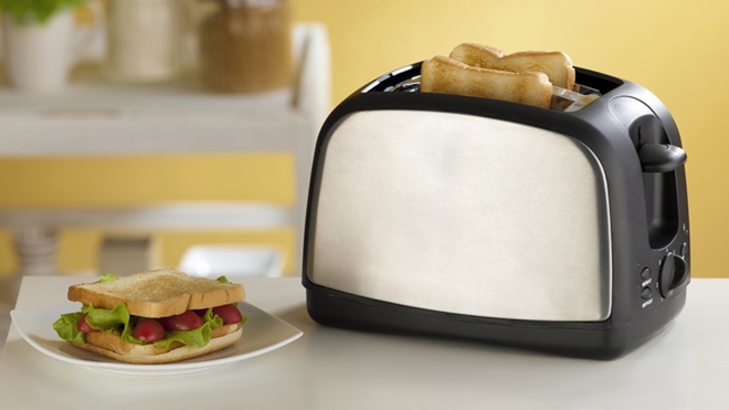 Em primeiro plano, sobre uma mesa, uma torradeira de cor preta e prateada exibe duas fatias de pão torradas e é ladeada por um prato com uma sanduíche de pão torrado, alface e tomate.