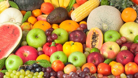 Detalhe de um conjunto variado e colorido de frutos, legumes e leguminosas, que incui: melão, melancia, meloa, maçãs, laranjas, uma maçaroca de milho, uvas, pimentos e tomates, entre outros.