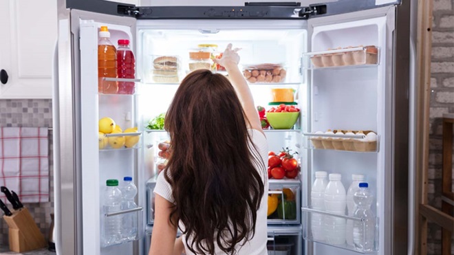 Mulher de frente para um frigorífico americano com as duas portas abertas lado a lado e escolher um produto na prateleira de cima 