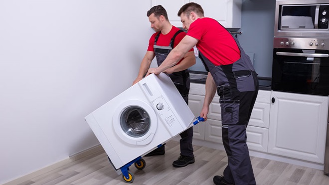 Dois trabalhadores carregam uma máquina de lavar roupa num trolley, numa cozinha particular - alusão a devolução de eletrodoméstico avariado