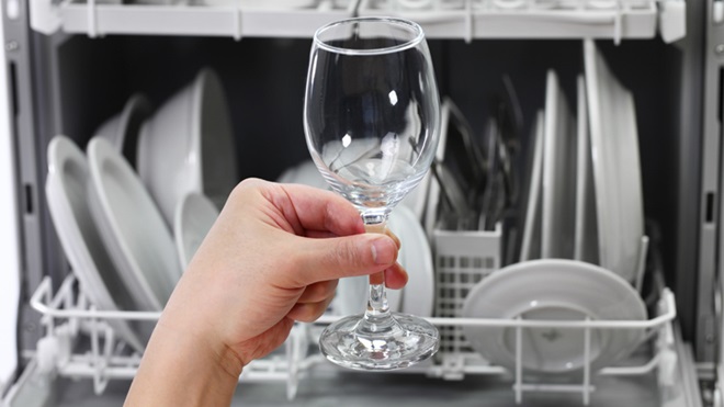 Como fundo da imagem, o tabuleiro superior de uma máquina de lavar loiça com loiça lavada no interior. Em primeiro plano, uma mão a agarrar um copo de água grande, também limpo.