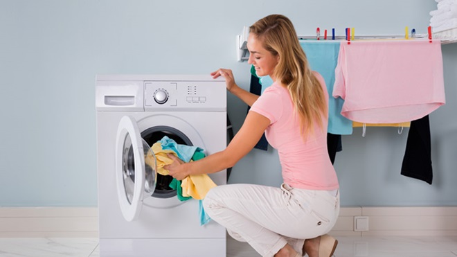 Mulher sorridentes de calças brancas e t-shirt cor de rosa coloca roupa dentro da máquina de secar.