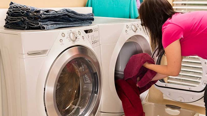 Senhora a tirar roupa da máquina de secar, instalada ao lado de uma máquina de lavar sobre a qual estão calças já dobradas e prontas a arrumar