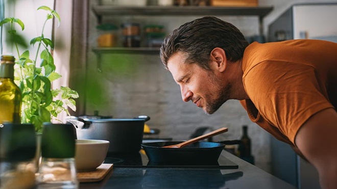 Homem com ar deliciado a cheirar o que está a ser cozinhado numa placa de indução. Cozinha moderna.