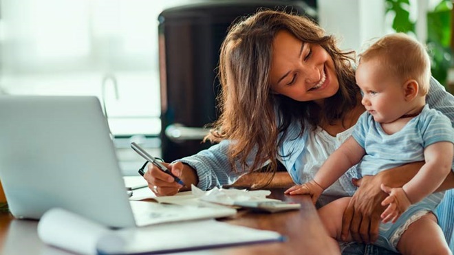 Mulher em frente ao computador em casa, segura o bebé ao colo, com ar sorridente