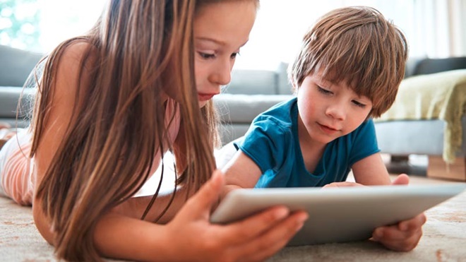Duas crianças, rapaz e rapariga, em casa, deitados no chão, consultam um tablet