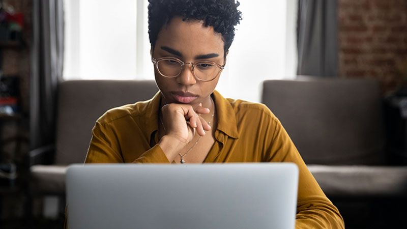 Pessoa sentada frente ao portátil, com postura pensativa e mão junto ao rosto, transmitindo concentração enquanto trabalha numa sala iluminada pela janela ao fundo.