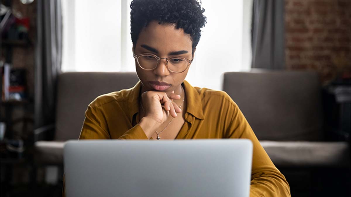 Pessoa sentada frente ao portátil, com postura pensativa e mão junto ao rosto, transmitindo concentração enquanto trabalha numa sala iluminada pela janela ao fundo.