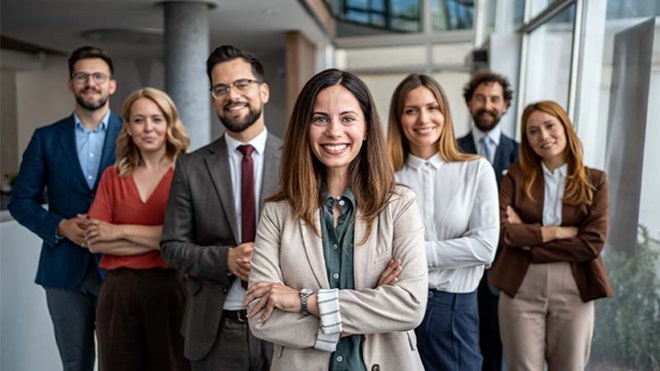 Grupo de sete pessoas alinhadas num espaço de escritório, com a figura da frente de braços cruzados e várias outras atrás, todas vestidas de forma profissional
