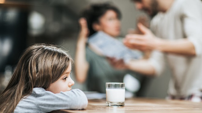 Criança triste sentada à mesa em frente de copo de leite, casal ao fundo a discutir