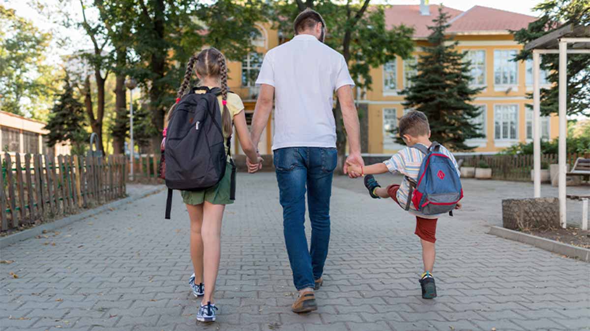 Uma pessoa adulta e duas crianças caminham num passeio arborizado em direção a um edifício escolar, de mãos dadas, com mochilas às costas e luz de manhã ao fundo.