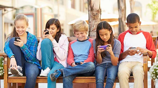 grupo de ciranças sentadas num banco no exterior, todas a olhar para o ecrã do telemóvel