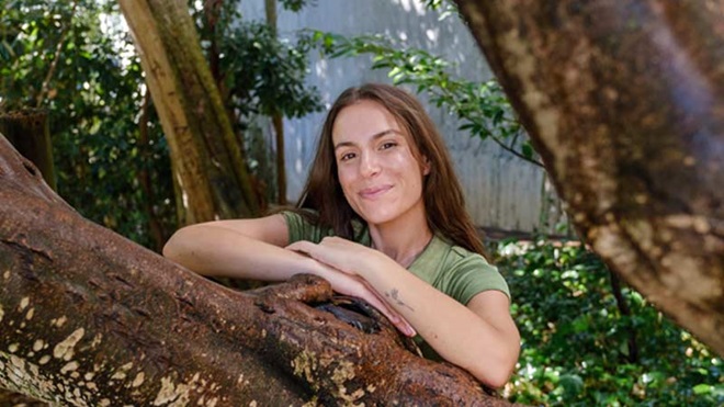 Sofia Manuel, a tripeirinha, mulher morena de cabelos compridos com braços cruzados sobre uma árvore a sorrir