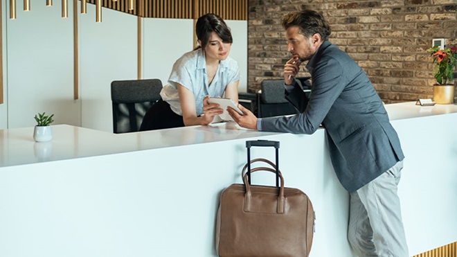 Cliente em lobby de hotel com mala de viagem a tirar dúvidas com rececionista