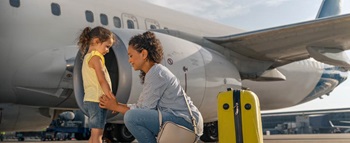 Uma mãe dá a mão à filha pequena, junto a um avião comercial, com mala de viagem amarela ao lado